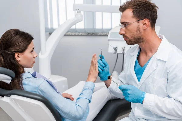 Bonito barbudo dentista segurando instrumentos dentários e olhando para assustado mulher gestos na clínica — Stock Photo