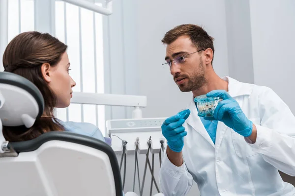 Foco seletivo de mulher atraente olhando para dentista bonito em óculos segurando modelo de dente — Fotografia de Stock