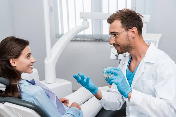 Dentista alegre em óculos segurando modelo de dente e olhando para a mulher feliz — Fotografia de Stock