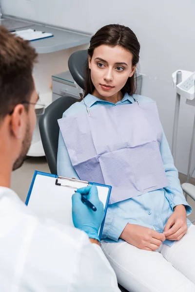 Selective focus of attractive patient looking at dentist writing diagnosis — Stock Photo