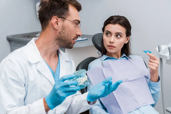 Messa a fuoco selettiva di bel dentista in occhiali gesticolando vicino alla donna con fermo — Foto stock