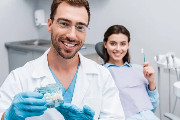 Селективный фокус счастливого стоматолога в очках с моделью зубов рядом с веселым пациентом с зубной щеткой — стоковое фото