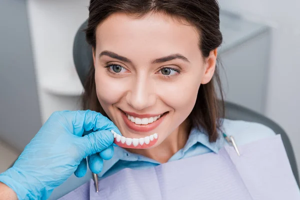 Vista recortada de dentista sosteniendo prótesis cerca de chica feliz en clínica dental - foto de stock