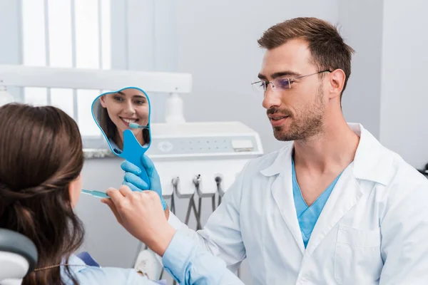 Selective focus of girl brushing teeth and looking at mirror near handsome dentist — Stock Photo