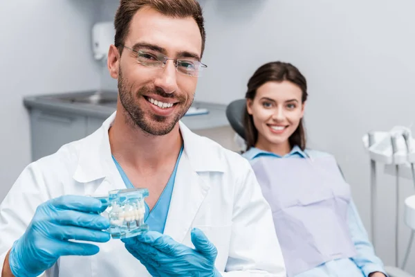 Селективный фокус счастливого бородатого стоматолога, держащего модель зубов рядом с счастливой девушкой — стоковое фото