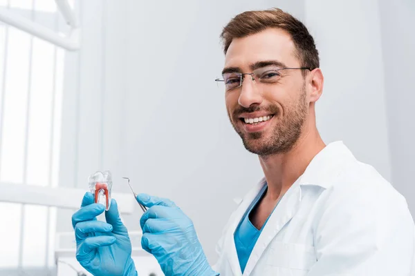 Красивый бородатый стоматолог с зубным инструментом и зубной моделью во время улыбки в клинике — стоковое фото