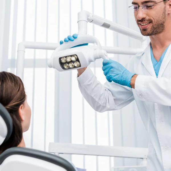 Selektiver Fokus des hübschen Zahnarztes in Latexhandschuhen, die medizinische Lampe in der Nähe des Patienten berühren — Stockfoto