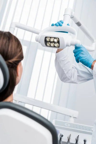 Обрезанный вид стоматолога в латексных перчатках касающихся медицинской лампы рядом с пациентом — стоковое фото