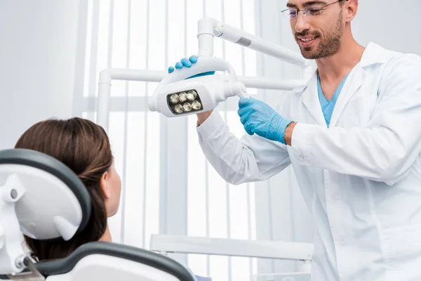 Selective focus of handsome dentist in blue latex gloves touching medical lamp near patient — Stock Photo