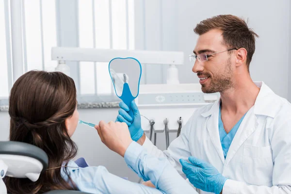 Enfoque selectivo del alegre dentista sosteniendo el espejo cerca de los dientes de cepillado del paciente - foto de stock