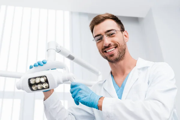 Vista de ángulo bajo de dentista alegre en gafas tocando lámpara médica y mirando a la cámara - foto de stock