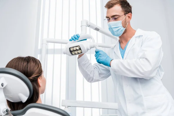 Вибірковий фокус стоматолога в медичній масці і синіх латексних рукавичках, що торкаються медичної лампи біля пацієнта — стокове фото