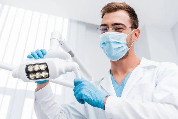 Vista de ángulo bajo del dentista en gafas y máscara médica tocando lámpara médica y mirando a la cámara - foto de stock