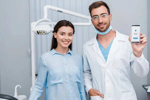 KYIV, UCRANIA - 10 de julio de 2019: niña alegre parada cerca del dentista con la mano en el bolsillo sosteniendo el teléfono inteligente con la aplicación de mensajero en la pantalla - foto de stock