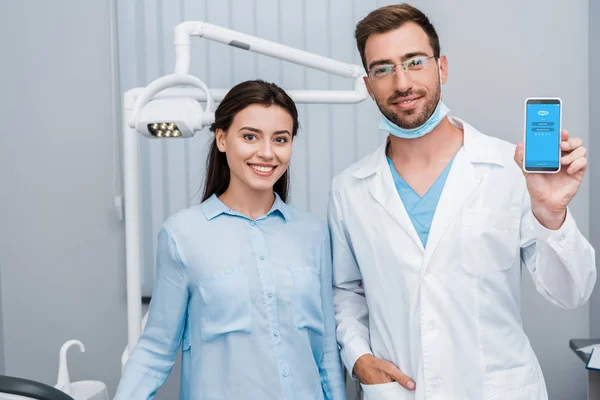 KYIV, UKRAINE - 10 JUILLET 2019 : joyeuse fille debout près du dentiste avec la main dans la poche tenant smartphone avec application skype à l'écran — Photo de stock