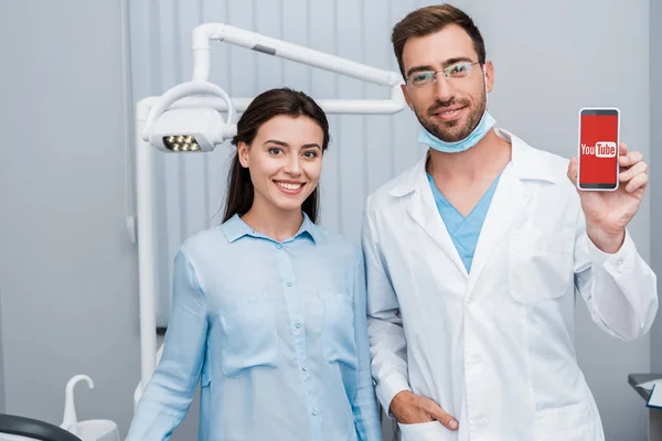 KYIV, UCRANIA - 10 de julio de 2019: chica alegre parada cerca del dentista con la mano en el bolsillo sosteniendo el teléfono inteligente con la aplicación de youtube en la pantalla - foto de stock