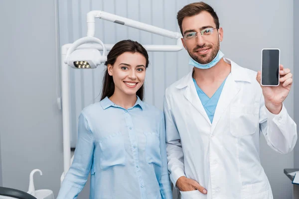 Fille gaie debout près du dentiste avec la main dans la poche tenant smartphone avec écran blanc — Photo de stock