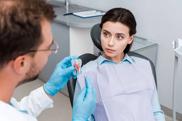 Messa a fuoco selettiva della giovane donna guardando dentista in guanti di lattice con modello di dente e strumento dentale — Foto stock