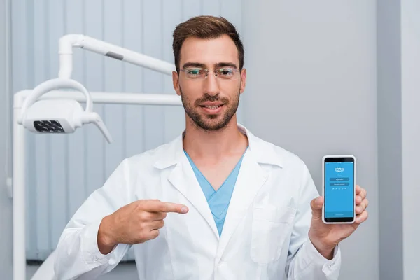KYIV, UKRAINE - JULY 10, 2019: handsome bearded man in white coat pointing with finger at smartphone with skype app on screen — Stock Photo