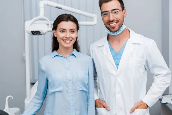 Весела дівчина посміхається біля красивого стоматолога, що стоїть з руками в кишенях — стокове фото