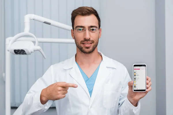 KYIV, UKRAINE - JULY 10, 2019: handsome bearded man in white coat pointing with finger at smartphone with apple music app on screen — Stock Photo
