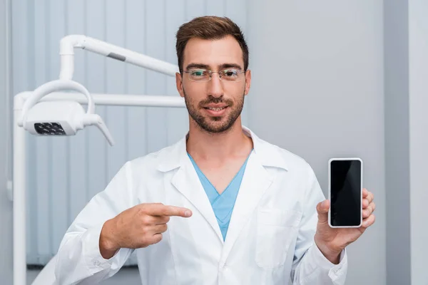 Hombre barbudo guapo con abrigo blanco apuntando con el dedo al teléfono inteligente con pantalla en blanco - foto de stock