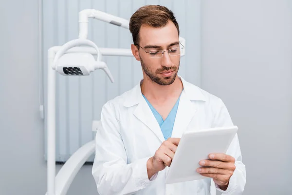 Hombre guapo de capa blanca y gafas usando tableta digital en la clínica - foto de stock