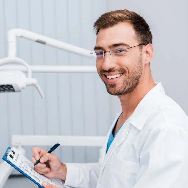 Homme barbu heureux en manteau blanc et lunettes tenant stylo et presse-papiers dans la clinique dentaire — Photo de stock