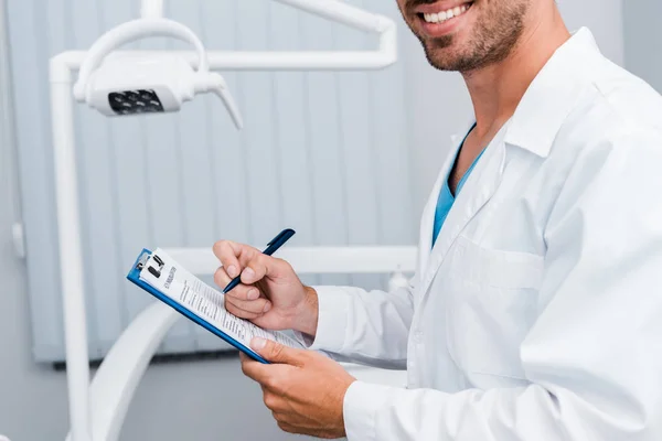 Vista recortada del hombre barbudo en blanco abrigo celebración pluma y portapapeles en clínica dental - foto de stock