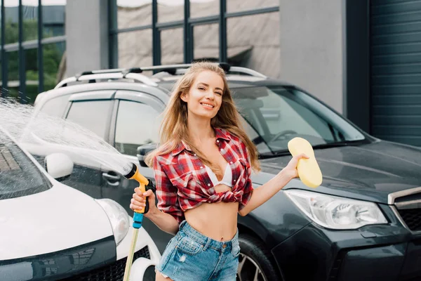 Alegre chica sosteniendo presión lavadora y esponja cerca de coches - foto de stock