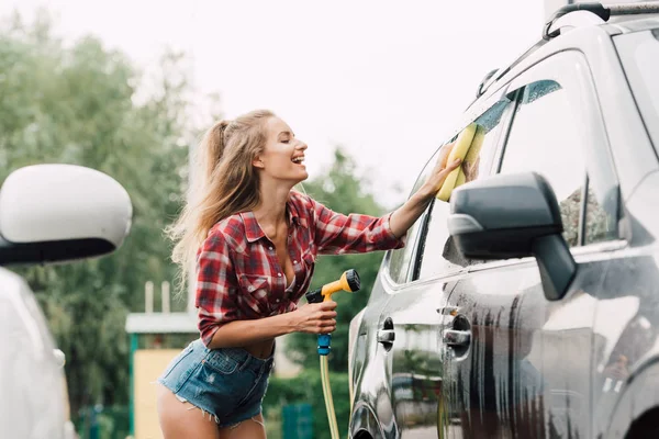 Chica atractiva sonriendo mientras se lava el coche con esponja - foto de stock