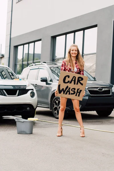 Ragazza positiva in piedi e in possesso di cartone con iscrizione di lavaggio auto vicino alle auto — Foto stock