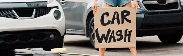 Colpo panoramico di ragazza in piedi e tenendo cartello di cartone con lettere di lavaggio auto vicino alle auto — Foto stock