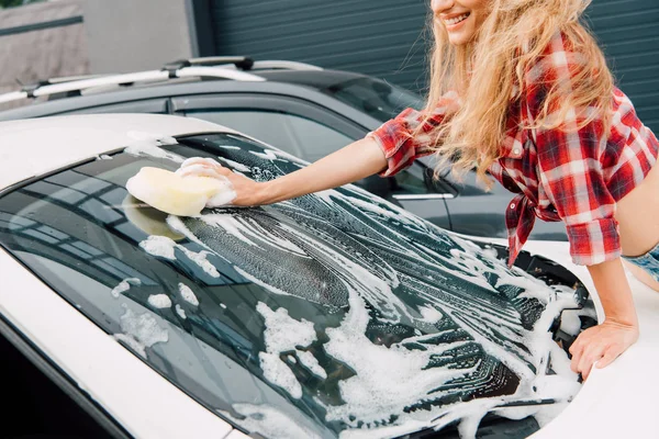 Cropped view of happy woman washing windshield of car with sponge and foam — Stock Photo