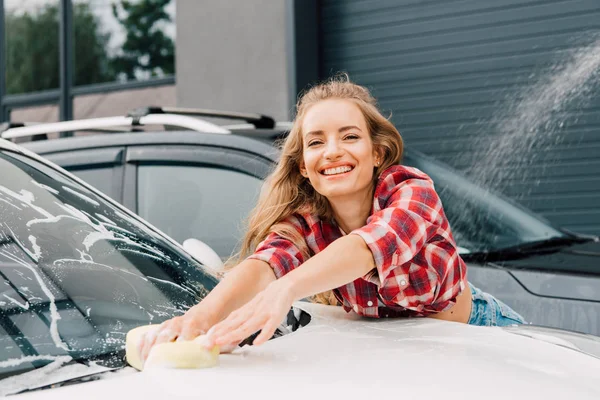 Mujer positiva lavando parabrisas de coche con esponja y espuma - foto de stock