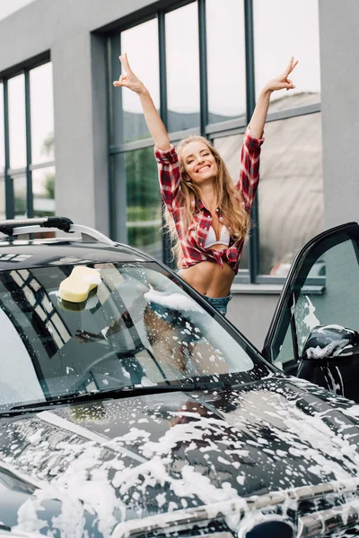 Glückliches Mädchen zeigt Friedenszeichen in der Nähe von nassem Auto mit Schaum und Gebäude — Stockfoto