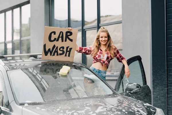 Junge Frau hält Karton mit Waschstraßen-Schriftzug in der Hand und steht neben Auto — Stockfoto
