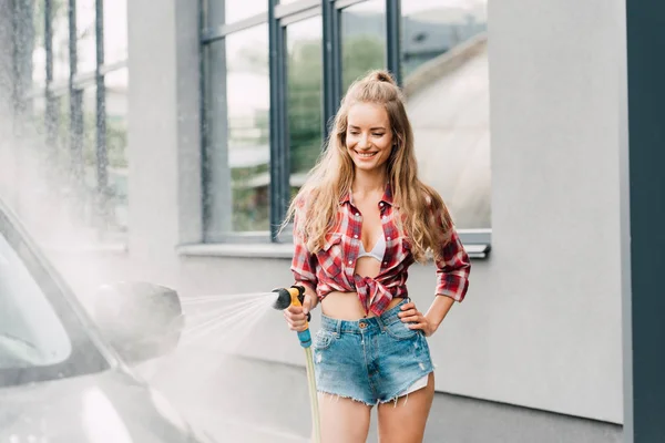 Happy girl standing with hand on hip and washing car near building — Stock Photo