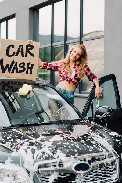 Fröhliche junge Frau hält Karton mit Autowäsche-Schriftzug in der Hand und steht neben Auto — Stockfoto