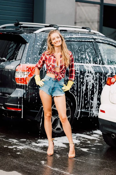 Fröhliches Mädchen, das mit den Händen auf Hüften in der Nähe von Autos in einer Waschanlage steht — Stockfoto