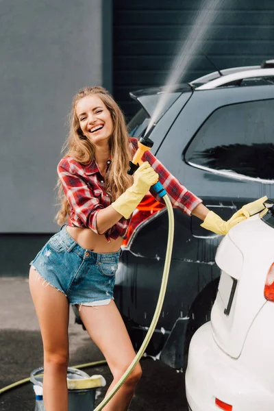 Alegre chica sosteniendo presión lavadora y sonriendo cerca de autos - foto de stock