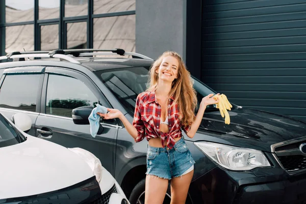 Mulher alegre segurando pano azul e luvas de látex amarelo perto de carros — Fotografia de Stock