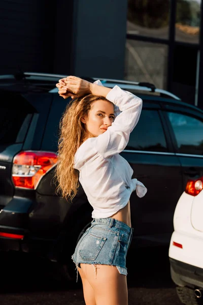 Attraktive junge Frau in blauen Jeanshosen blickt in die Kamera in der Nähe von Autos — Stockfoto