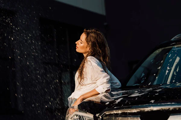 Привлекательная и мокрая девушка, стоящая возле черного роскошного автомобиля — стоковое фото