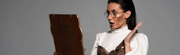Plano panorámico de mujer steampunk impactada en gafas mirando la pantalla del ordenador portátil vintage aislado en gris - foto de stock