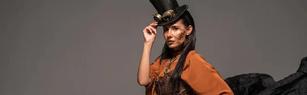 Panoramic shot of attractive steampunk woman in top hat with goggles looking at camera isolated on grey — Stock Photo