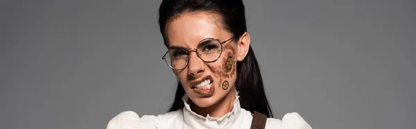 Panoramic shot of irritated steampunk woman in glasses with makeup looking at camera isolated on grey — Stock Photo