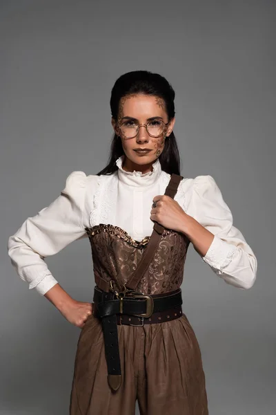 Vista frontal de la mujer steampunk con maquillaje de pie con la mano en la cadera aislado en gris - foto de stock