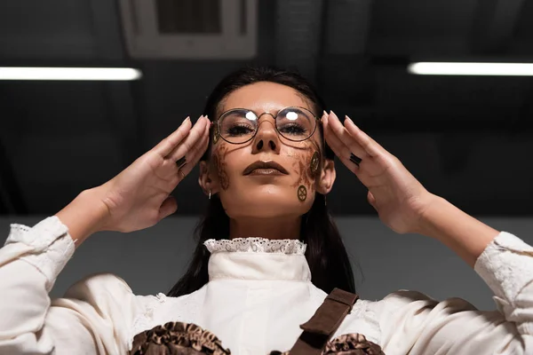 Low angle view of steampunk woman touching glasses and looking at camera — Stock Photo