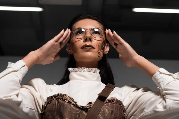Low angle view of steampunk woman touching glasses and looking up — Stock Photo